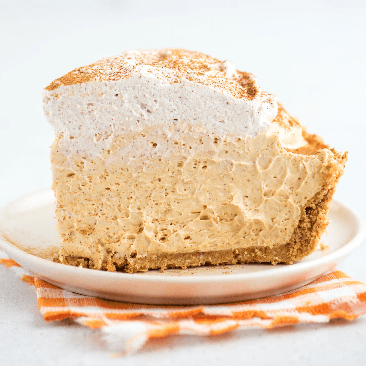 Questa torta di zucca senza cottura, veloce e facile, è leggera e cremosa - una deliziosa variante della classica torta di zucca. Un pezzo leggero e cremoso di torta di zucca senza cottura adagiato su un piatto da dessert, sopra un tovagliolo a quadretti arancioni.