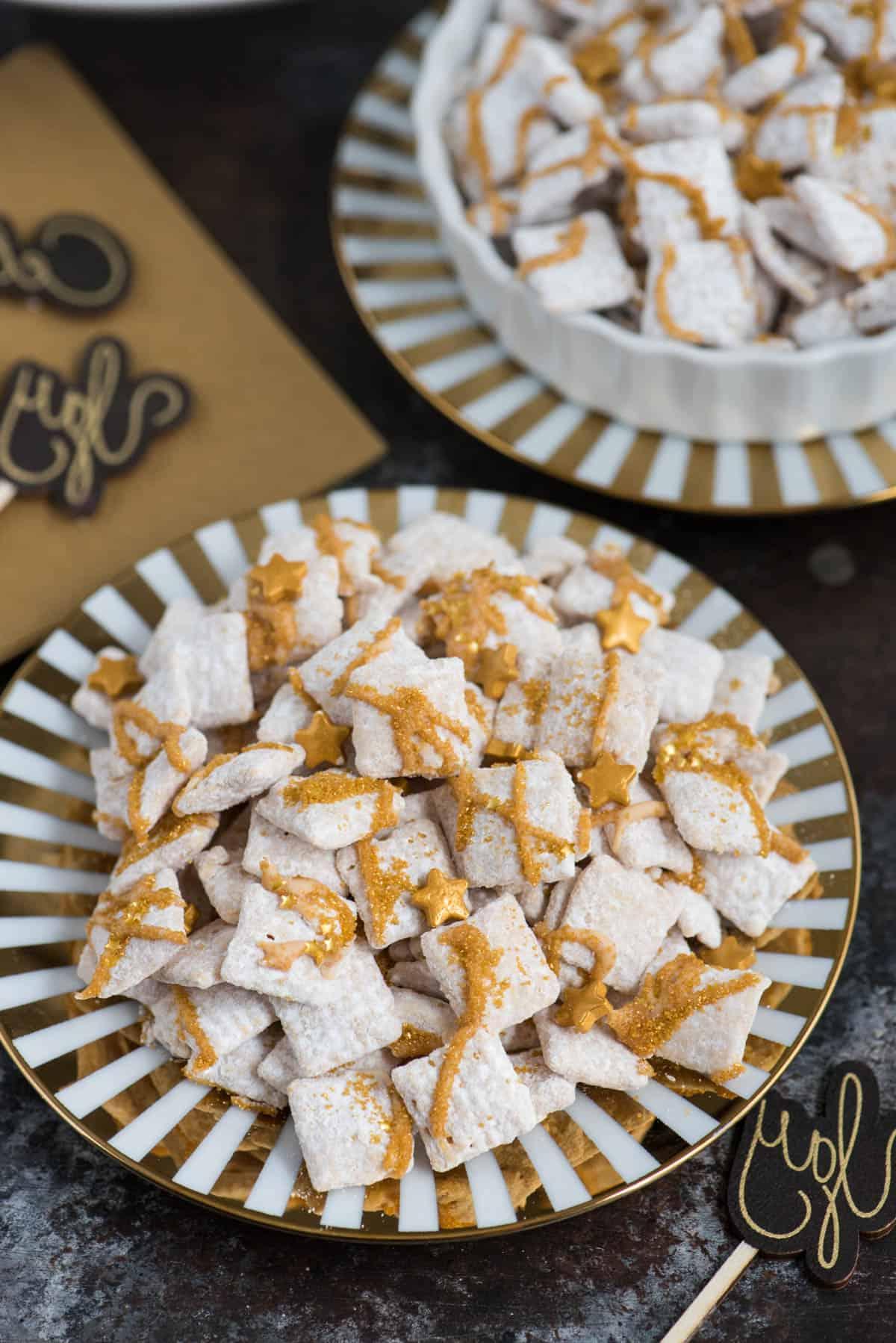 **Dolcetti Croccanti al Cioccolato Bianco di Capodanno** 2 new year’s eve white chocolate puppy chow with gold sprinkles and gold stars on a gold striped plate on dark black background