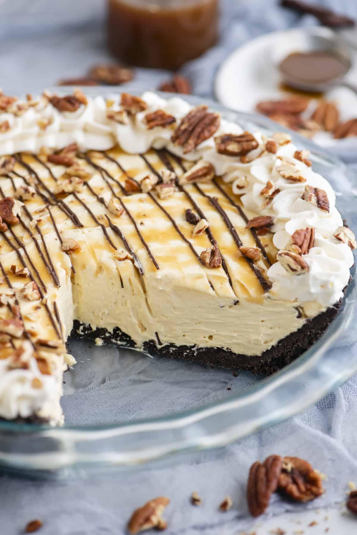 Torta di Tartaruga 2 Torta Tartaruga in un piatto di vetro, guarnita con caramello, cioccolato, panna montata e noci pecan, con noci pecan sparse intorno.