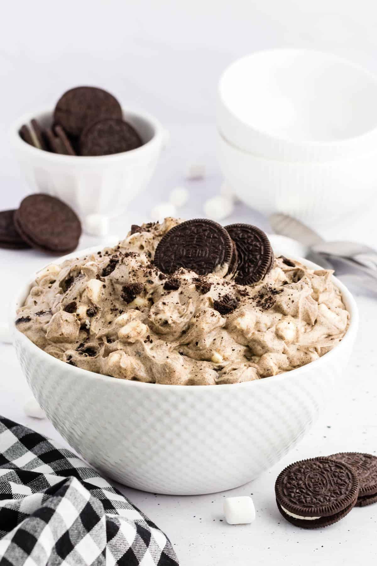 Ricetta Crema Soffice Oreo (Senza Formaggio Cremoso) 2 Fluff di Oreo in una ciotola bianca circondato da biscotti Oreo interi, mini marshmallow e un panno da cucina a scacchi bianchi e neri