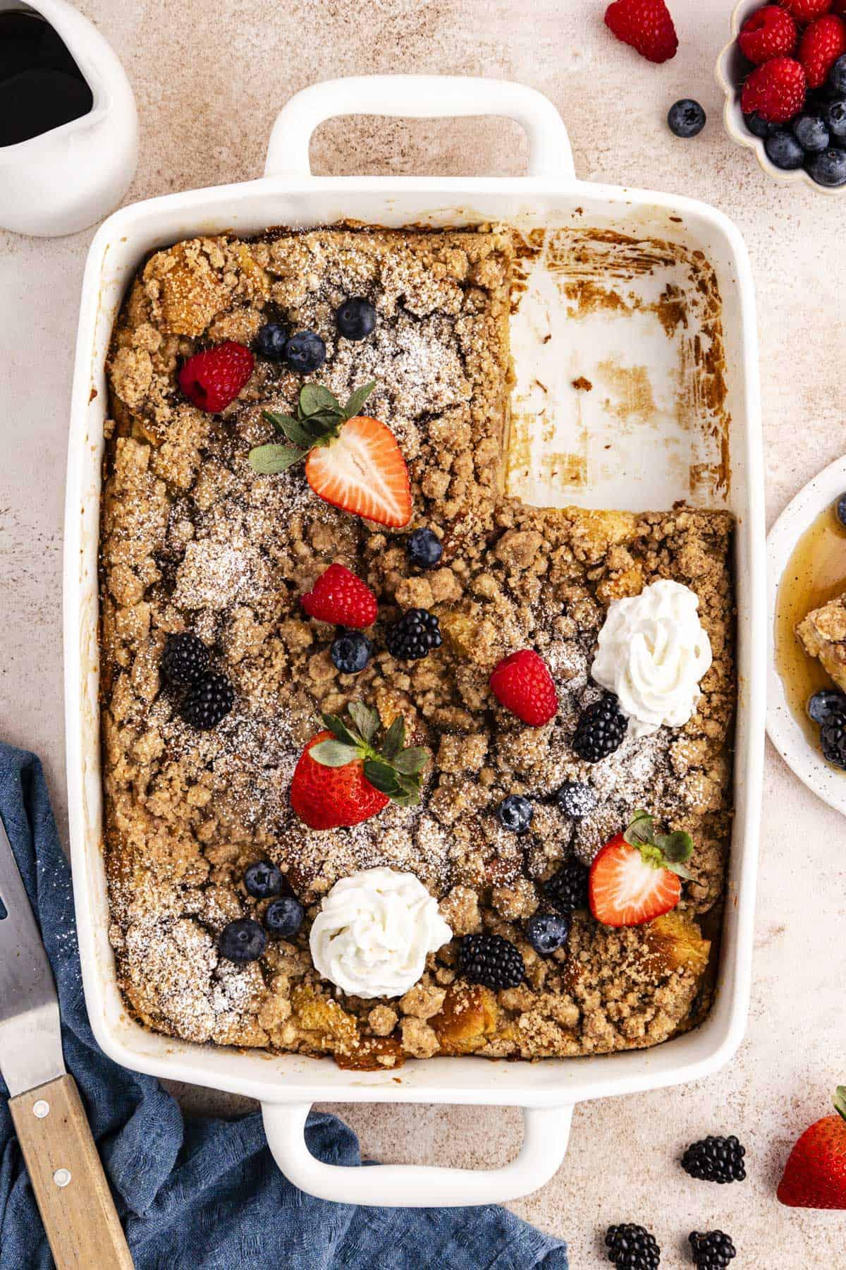 Migliore Ricetta per Budino di French Toast 2 Una teglia bianca piena di casseruola di French Toast con una fetta mancante, guarnita con frutti di bosco freschi, ciuffi di panna montata e una leggera spolverata di zucchero a velo, circondata da una ciotola di frutti di bosco, una ciotola di sciroppo, un piatto con una fetta e sciroppo, un canovaccio blu e una spatola.