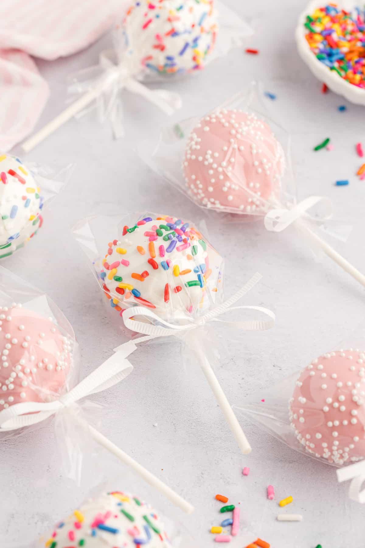 cake pops singolarmente incartati nella plastica con fiocchi bianchi, adagiati su una superficie grigio chiaro, alcuni rosa con codette bianche e alcuni bianchi con codette arcobaleno, con codette arcobaleno sparse intorno