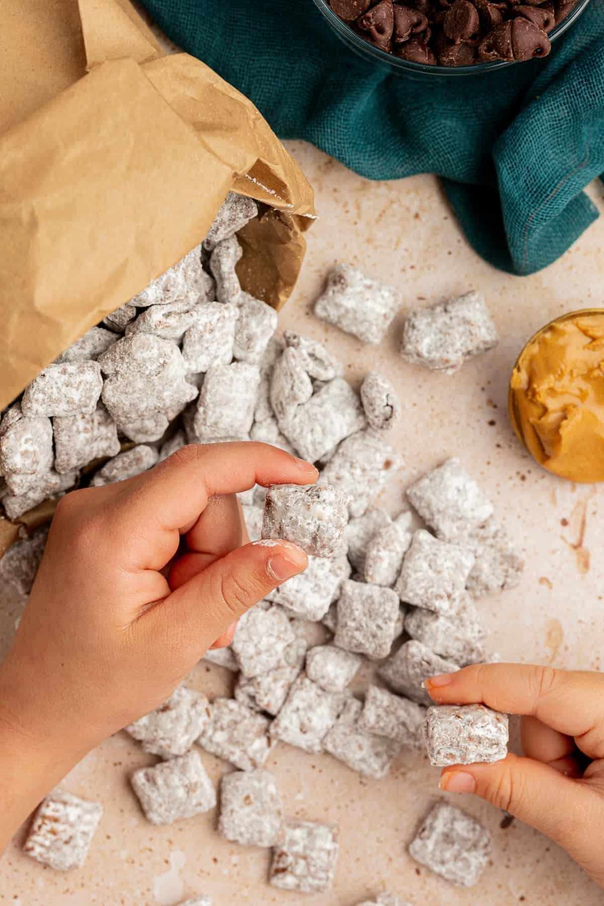 Una borsa marrone piena di puppy chow adagiata su un fianco con il puppy chow che si rovescia, due mani che tengono pezzi di puppy chow, una paletta di burro d'arachidi accanto e un asciugamano color ottanio scuro