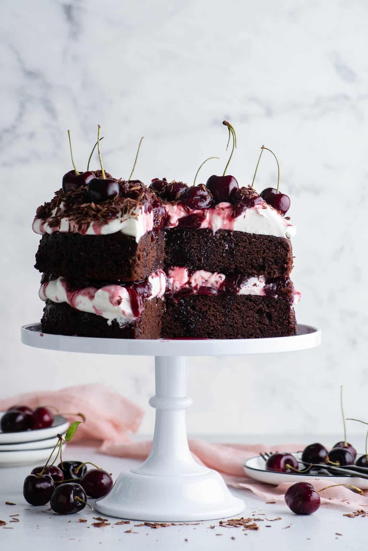 Torta Foresta Nera 2 Torta Foresta Nera su un'alzatina bianca con una grande fetta tagliata, con pile di piatti bianchi e ciliegie sparse intorno