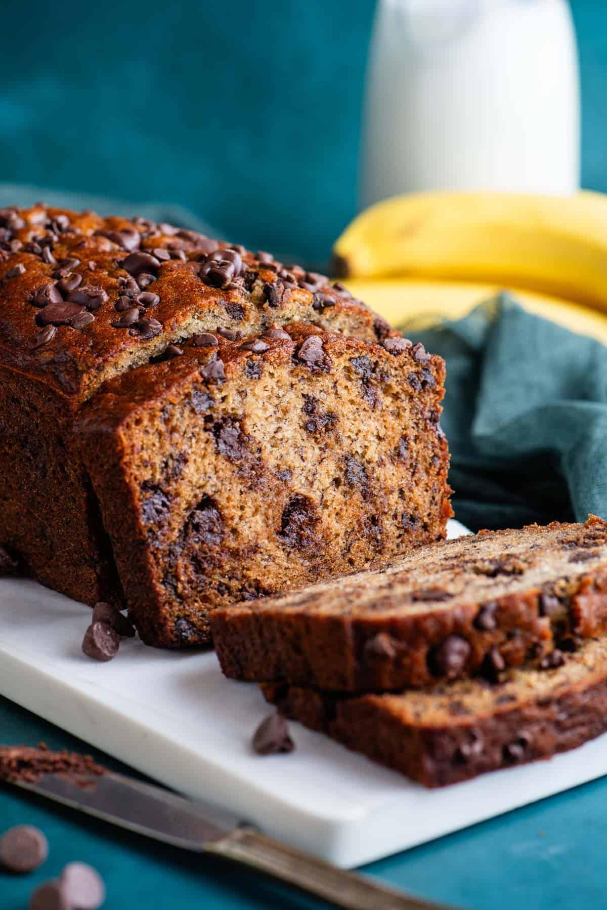 Il Miglior Pan di Banane con Gocce di Cioccolato 2 Una pagnotta di pane alle banane e gocce di cioccolato su un tagliere bianco con un asciugamano color ottanio scuro accanto, gocce di cioccolato sparse intorno, e banane intere e un bicchiere di latte sullo sfondo.