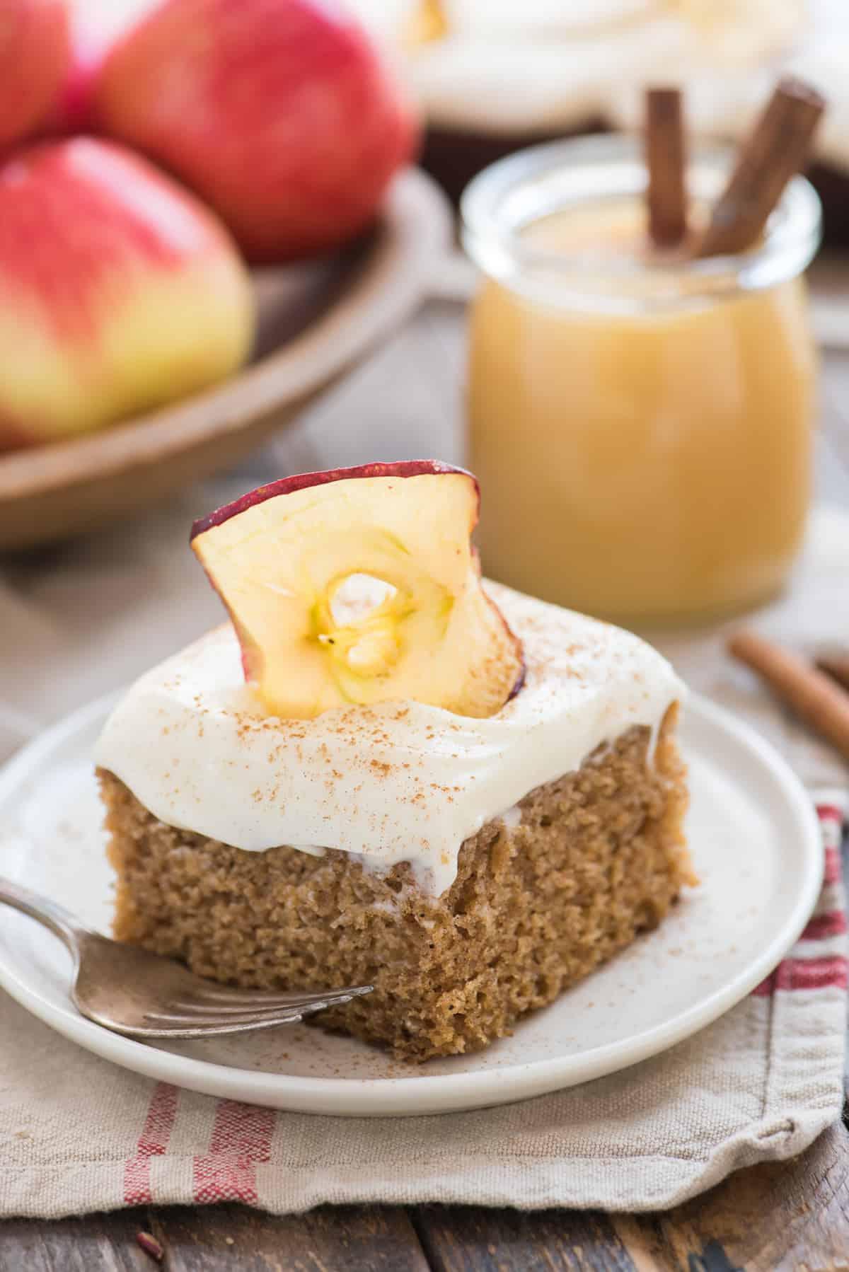 Torta Speziata alla Salsa di Mele 2 Fetta di torta di mele con glassa al formaggio cremoso su un piatto bianco con mele sullo sfondo