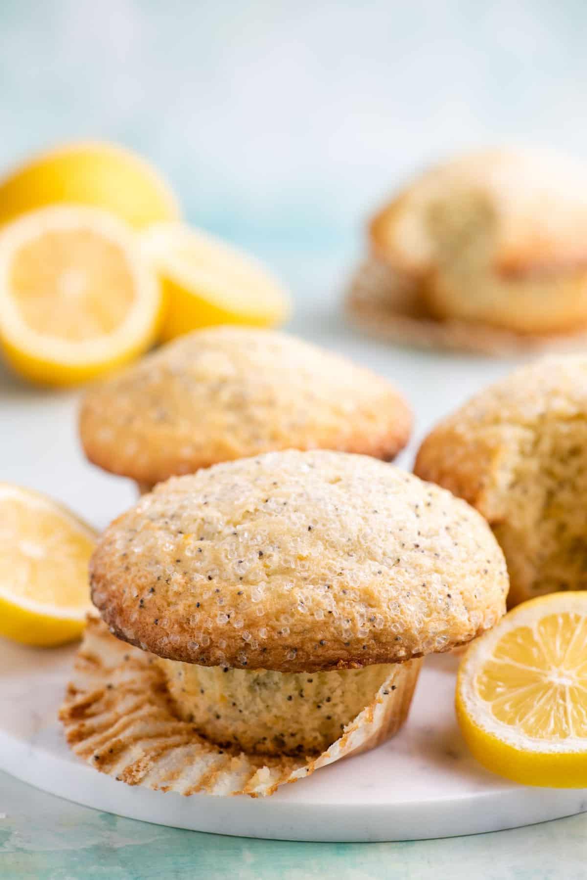 Migliori Muffin al Limone e Semi di Papavero 2 muffin al limone e semi di papavero su un piatto bianco con la carta parzialmente rimossa, circondato da altri muffin e fette di limone