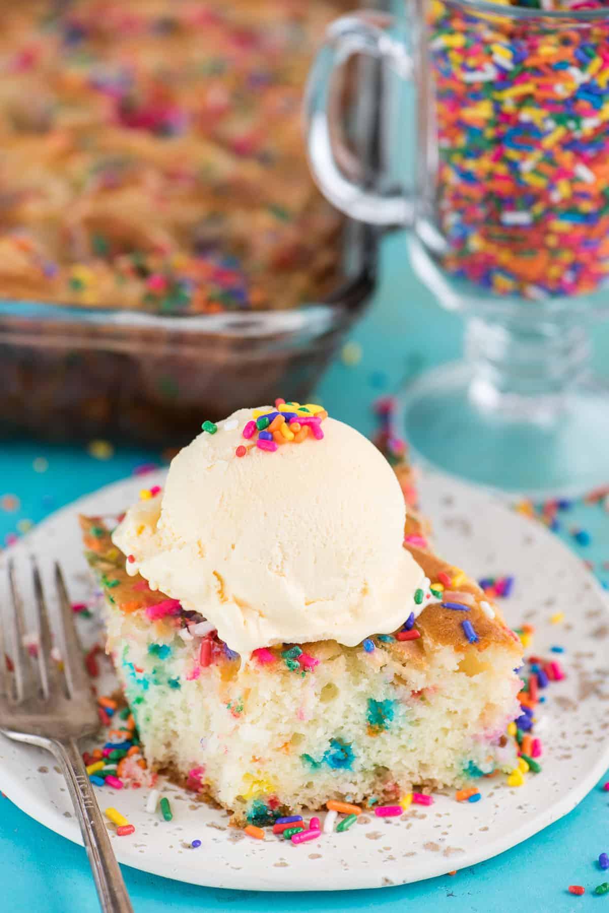 Questa torta terremoto funfetti è un mix per torta bianca arricchito con zuccherini e gocce di cioccolato funfetti. Lo strato di glassa al formaggio cremoso si fonde nella torta creando crepe e fessure in superficie. #earthquakecake #funfetticake #cake Fetta di torta terremoto funfetti con pallina di gelato alla vaniglia su piatto bianco su sfondo blu