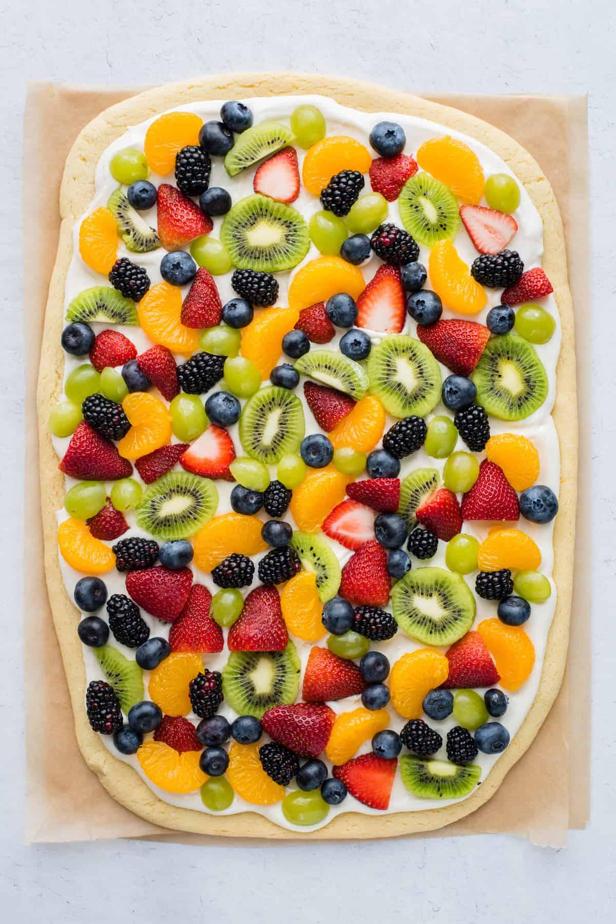 Una pizza di frutta intera con fragole, mirtilli, mandarini, kiwi e uva su carta da forno marrone