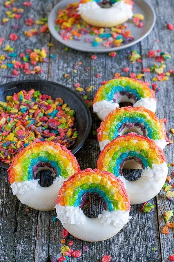 Ciambelle Arcobaleno - queste divertenti ciambelle sono fatte con cereali colorati, cioccolato bianco e cocco grattugiato! Non potrai fare a meno di queste deliziose ciambelle! Ciambelle Arcobaleno con cioccolato bianco, scaglie di cocco e cereali colorati su un tavolo di legno.