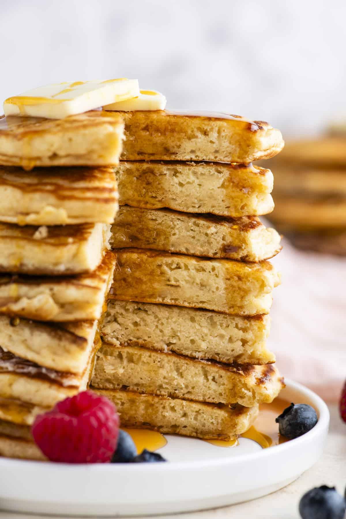 Pancakes Soffici Fatti in Casa 2 Una torre alta di pancake con una porzione triangolare tagliata, condita con sciroppo, sormontata da una fetta di burro e circondata da frutti di bosco freschi su un piatto bianco.