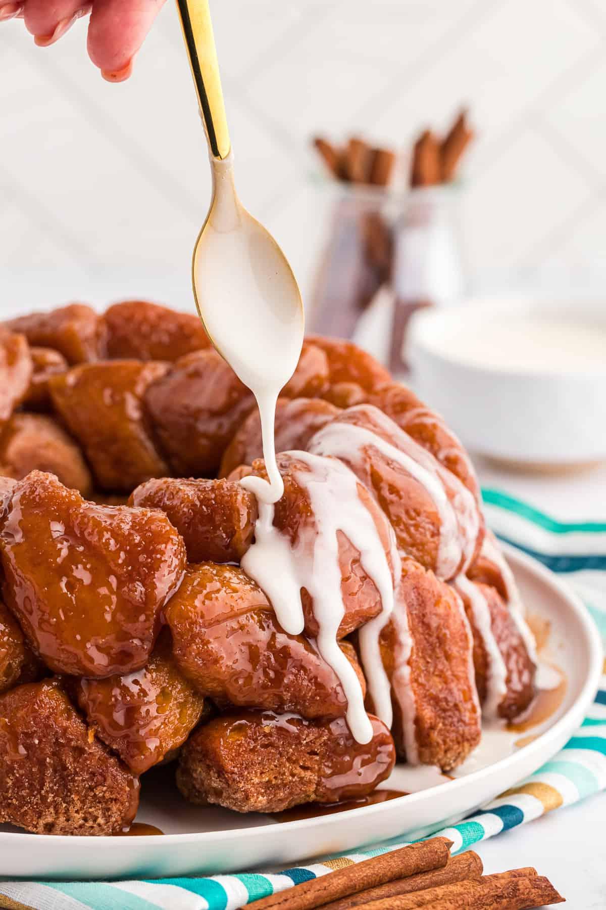 Pane delle scimmie di biscotti 2 Glassa versata su un piatto di Monkey Bread con un cucchiaio dorato, il piatto è posizionato su un telo a strisce turchese, blu e oro con bastoncini di cannella accanto