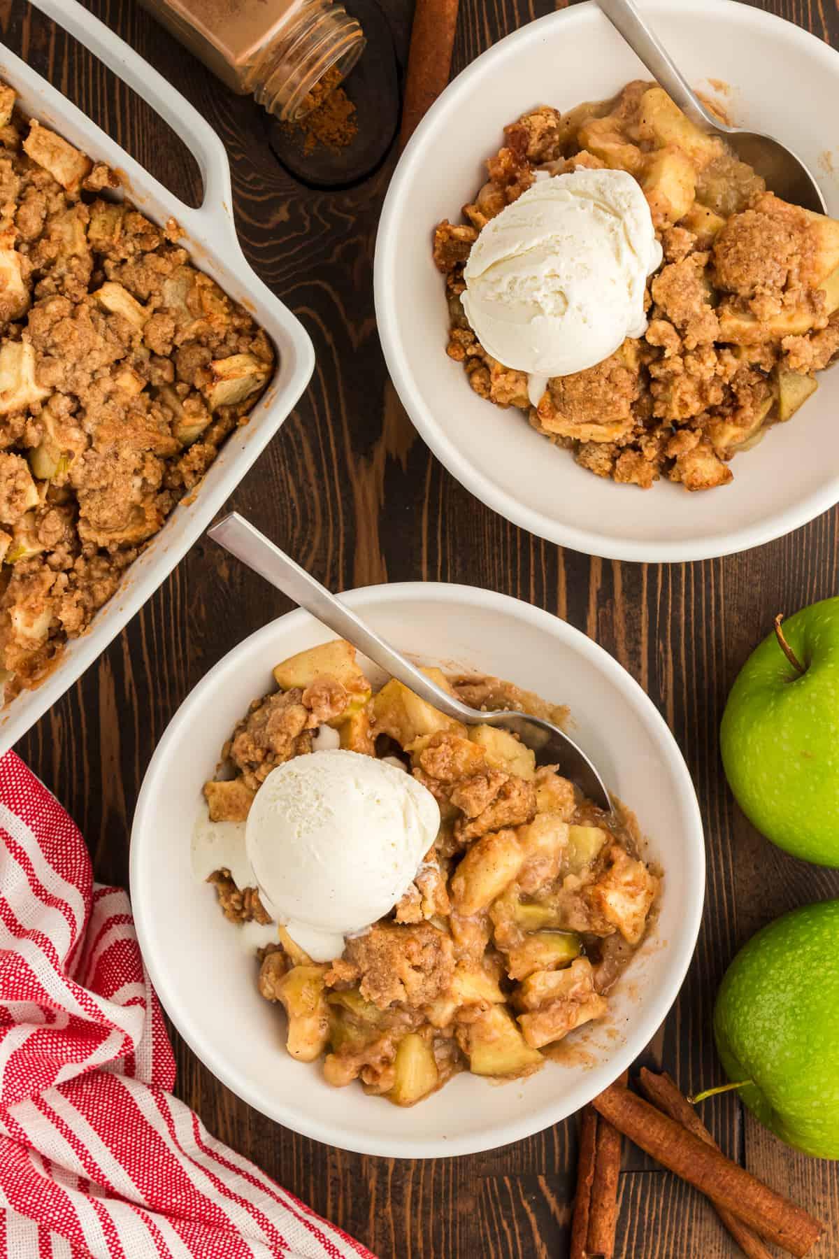 Ricetta Facile di Crumble alle Mele 2 Due ciotole di crumble di mele con una pallina di gelato alla vaniglia e un cucchiaio, accanto a una pirofila bianca piena di crumble di mele, tutto su una superficie di legno circondata da un canovaccio a righe rosse e bianche, due mele verdi, bastoncini di cannella e alcune spezie