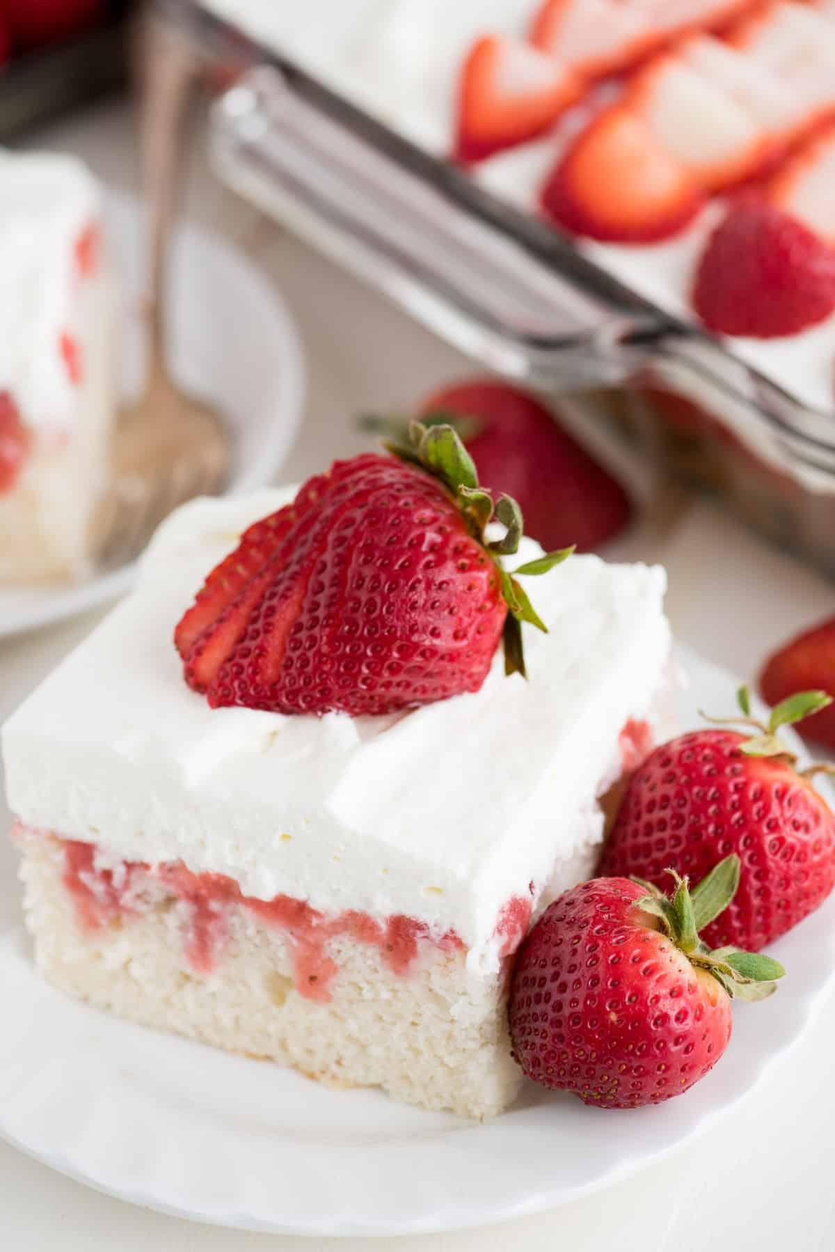 Poke Cake alle Fragole Fresche 2 Una fetta di torta poke alle fragole con salsa di fragole fatta in casa e copertura di panna, decorata con una fragola fresca. La torta è umida e invitante.