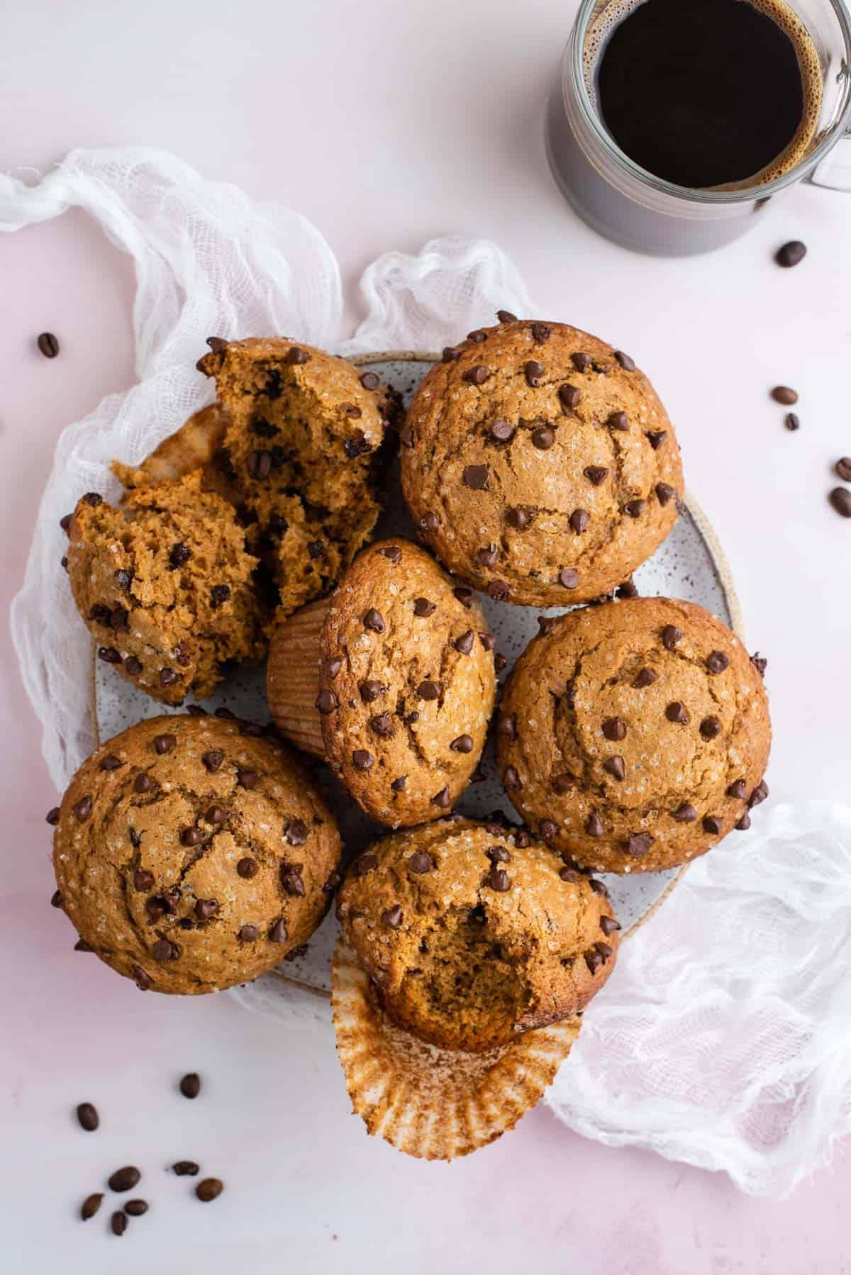 un piatto pieno di muffin al cappuccino con gocce di cioccolato su un telo di formaggio bianco con chicchi di caffè sparsi intorno e una tazza di caffè di vetro trasparente accanto