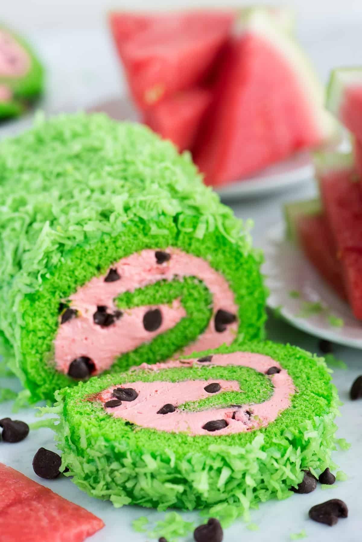 Una torta rotolo verde brillante con un ripieno rosa intenso e gocce di cioccolato che imitano i semi di anguria.