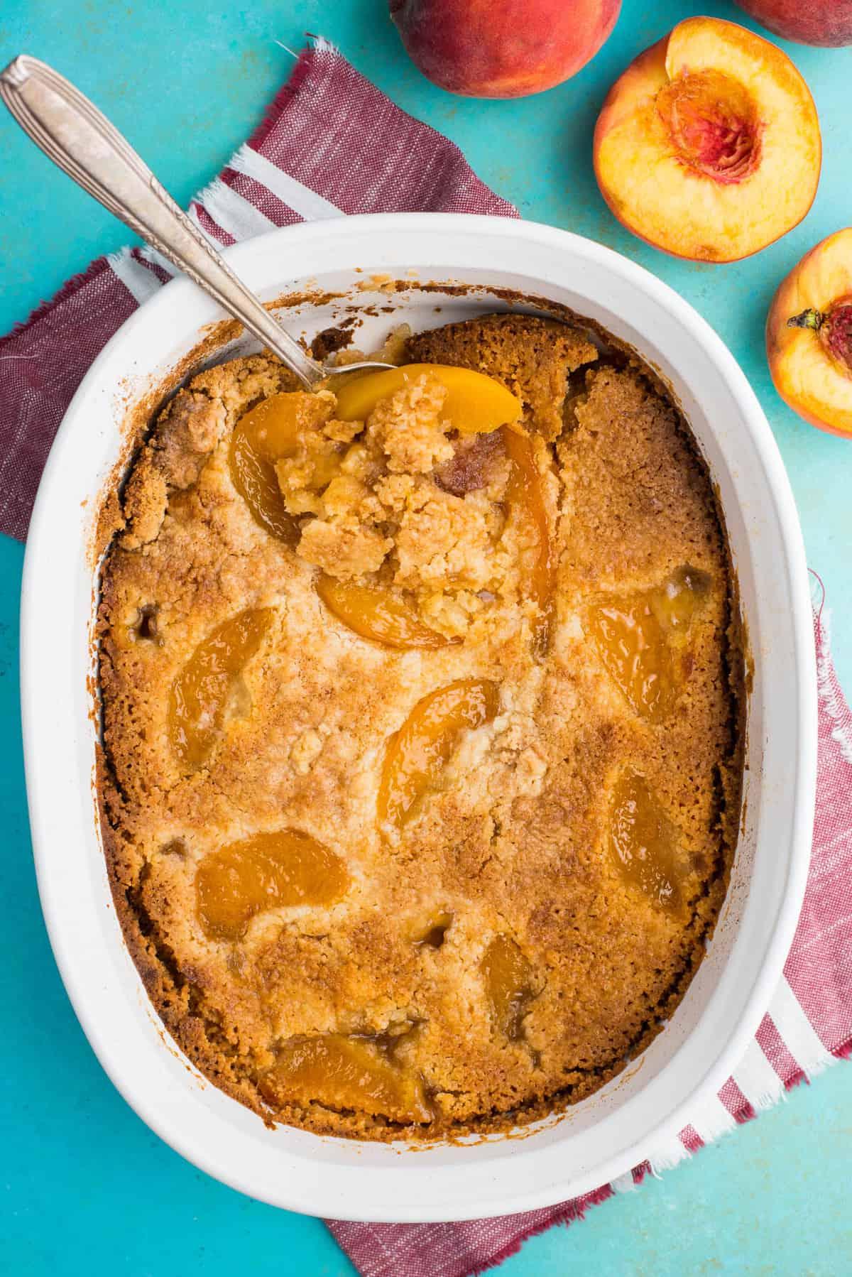 Torta Facile di Pesche con Preparato per Torta 2 torta di pesche dump cake con cucchiaio in teglia bianca su sfondo verde acqua