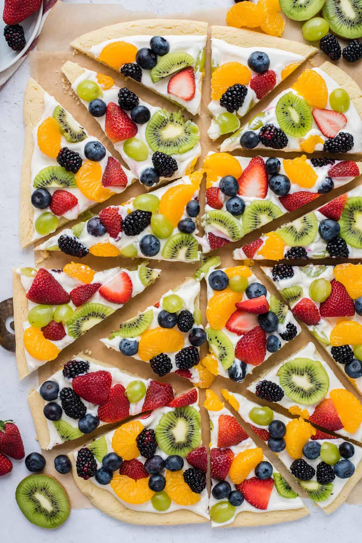 Una pizza di frutta intera tagliata a fette triangolari con fragole, mirtilli, mandarini, kiwi e uva su carta da forno marrone