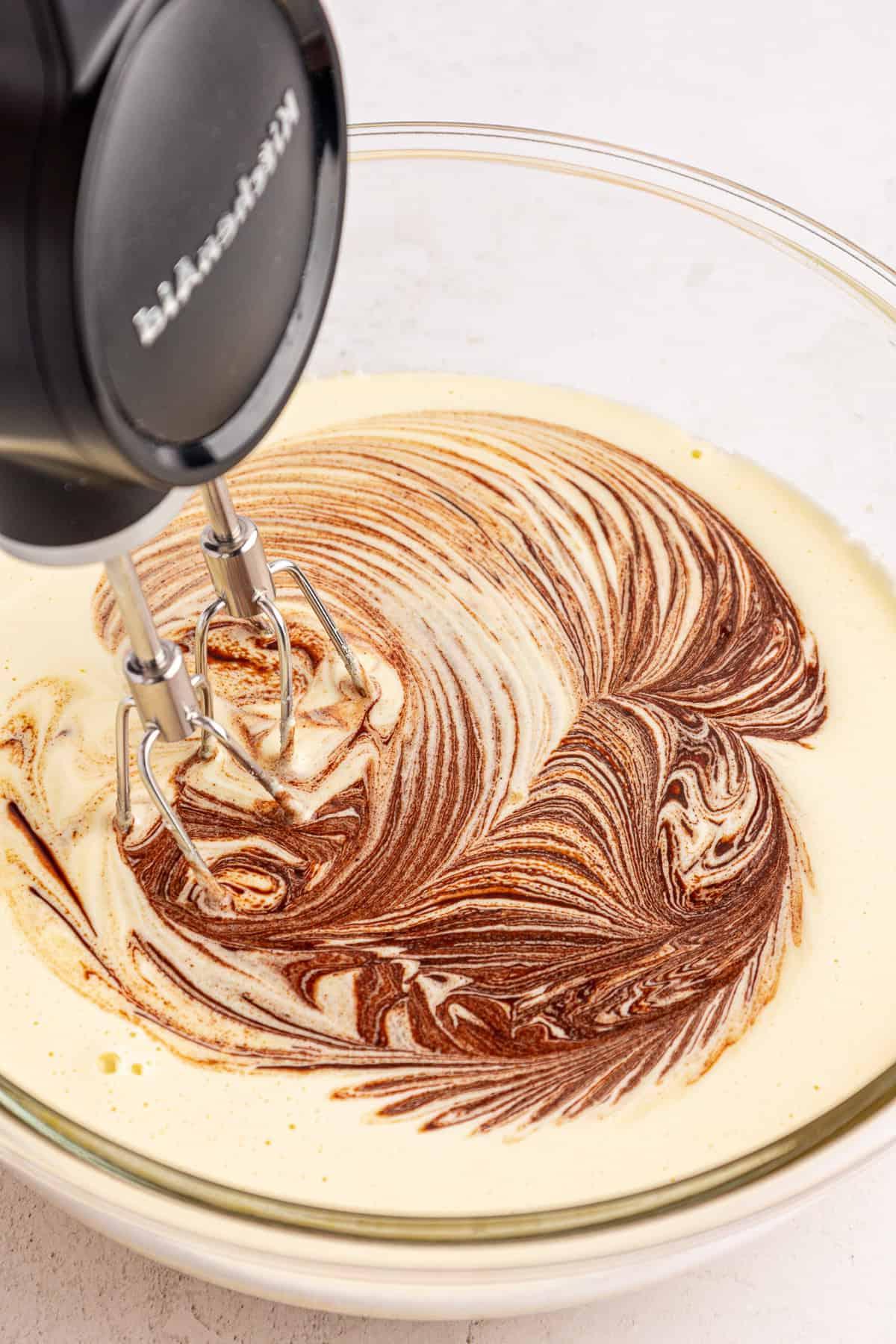 Una planetaria mescola un impasto giallo chiaro con spirali di cioccolato in una ciotola di vetro trasparente, creando un effetto marmorizzato.
