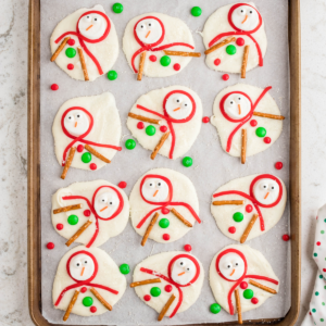 Una teglia con dodici biscotti pupazzo di neve sciolti ispirati al bark, ciascuno decorato con sciarpe di glassa rossa, braccia di bastoncini di pretzel, bottoni di caramella e faccine. M&Ms verdi e rossi sono sparsi sulla carta forno intorno ai biscotti.