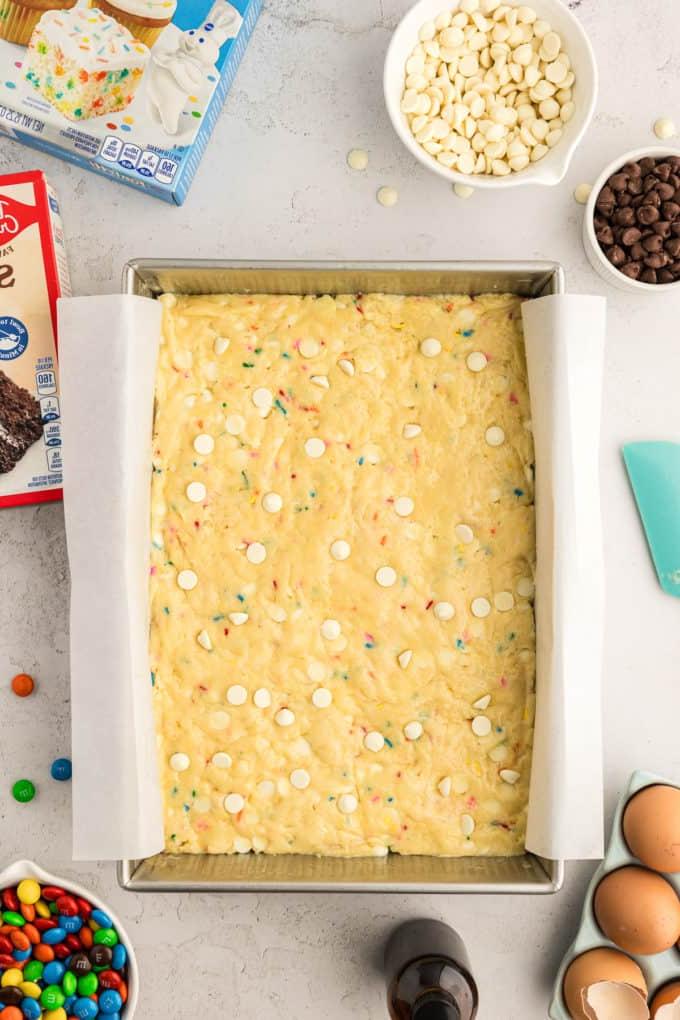 Barrette di biscotti da preparato per torte 11 a rectangular pan of funfetti cake bar batter surrounded by two boxes of cake mix, eggs, a bottle of vanilla, a bowl of M&Ms, a small bowl of chocolate chips and a larger bowl of white chocolate chips