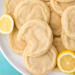 Ricetta Biscotti al Limone 10 Un piatto di biscotti al limone morbidi e ricoperti di zucchero, con un biscotto al limone morsicato. Fette di limone fresco sono posizionate accanto ai deliziosi biscotti al limone sul piatto.