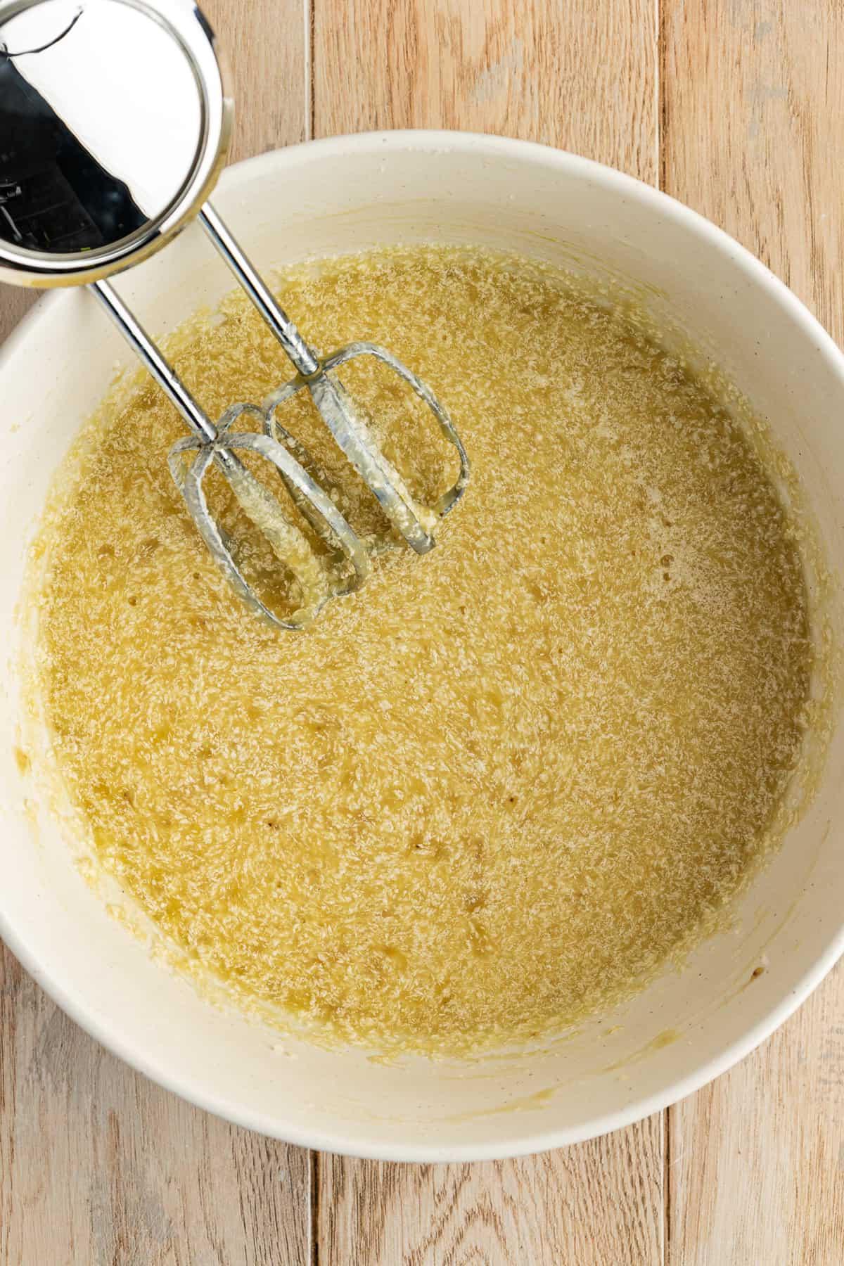 Ricetta Facile Torta alla Banana 11 Una ciotola bianca su una superficie di legno con un impasto di torta di banane e un mixer elettrico appoggiato all'interno della ciotola