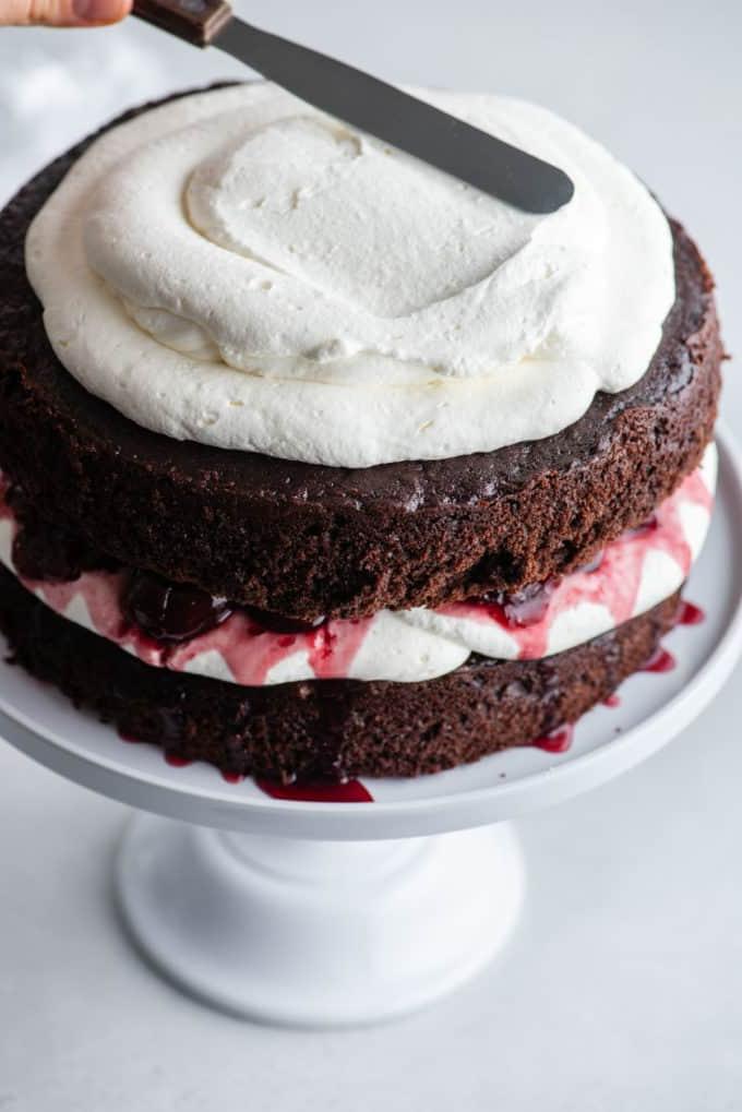 Torta Foresta Nera 11 Torta Foresta Nera su un'alzatina bianca con uno strato di torta al cioccolato, panna montata, ciliegie, più torta al cioccolato e panna montata che viene spalmata sopra