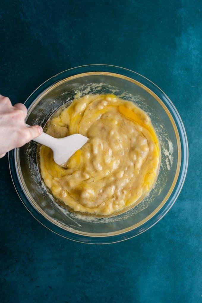Il Miglior Pan di Banane con Gocce di Cioccolato 11 Ingredienti liquidi per il pane alle banane e gocce di cioccolato che vengono mescolati con una spatola bianca in una ciotola di vetro trasparente su una superficie color ottanio scuro.