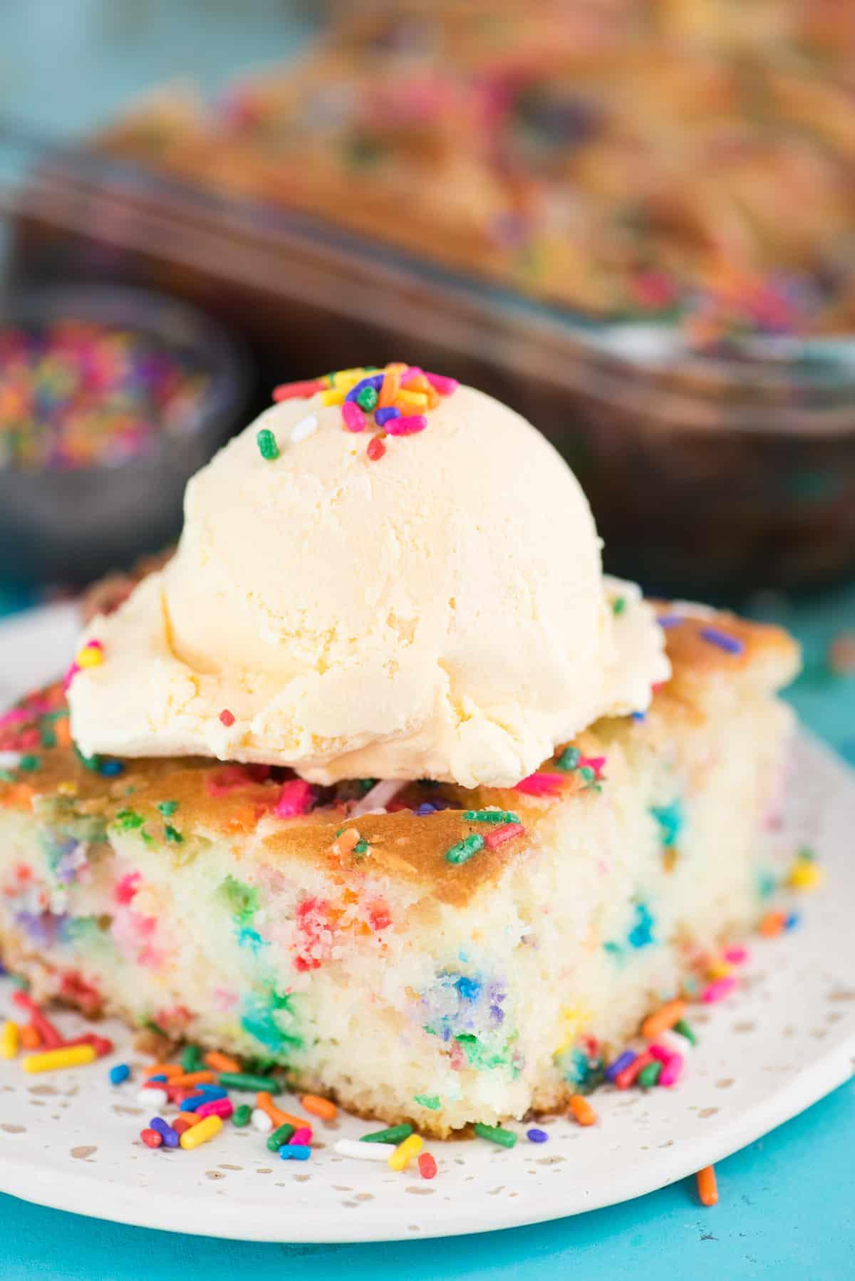 Questa torta terremoto funfetti è un mix per torta bianca arricchito con zuccherini e gocce di cioccolato funfetti. Lo strato di glassa al formaggio cremoso si fonde nella torta creando crepe e fessure in superficie. #earthquakecake #funfetticake #cake Fetta di torta terremoto funfetti con pallina di gelato alla vaniglia su piatto bianco su sfondo blu