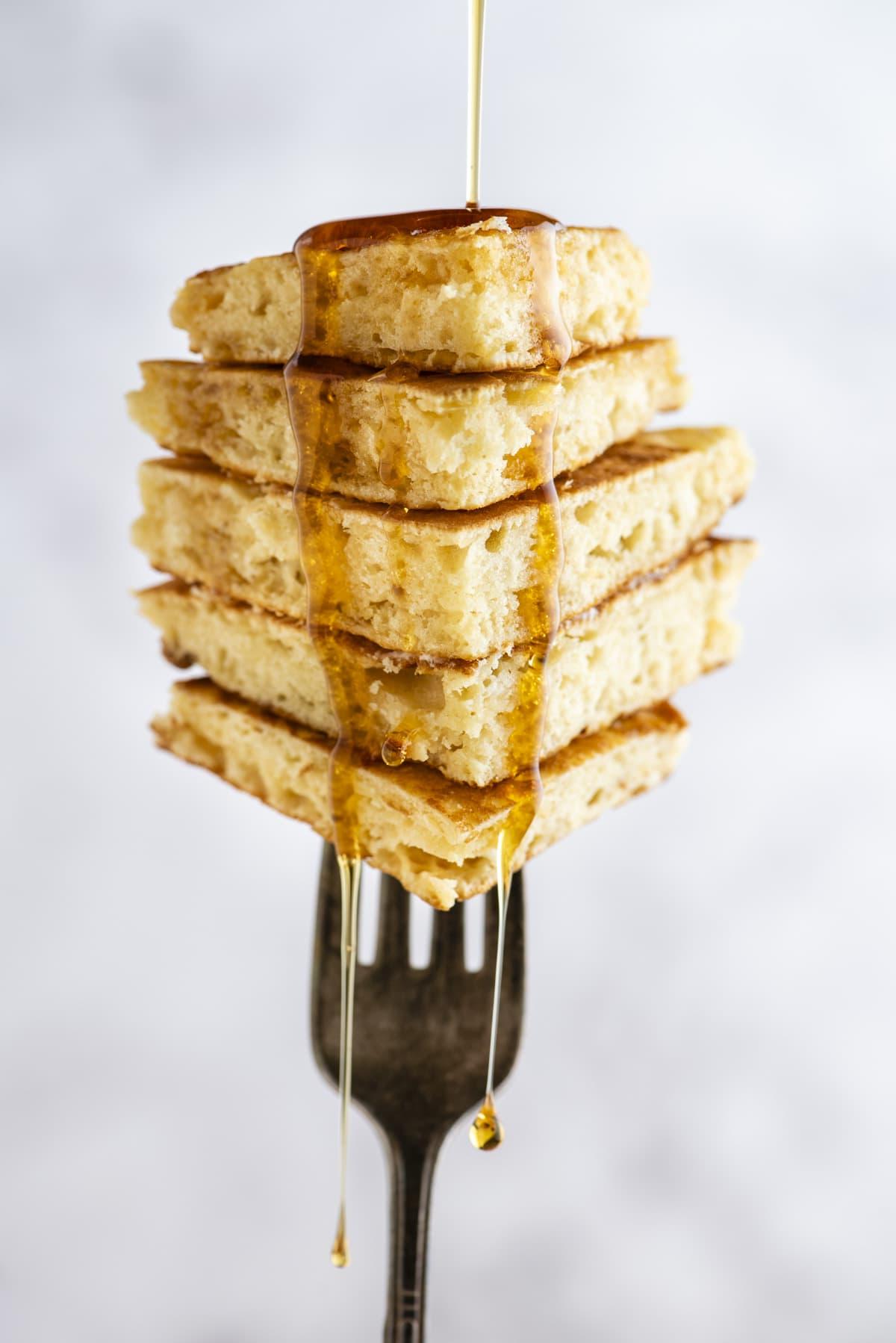 Pancakes Soffici Fatti in Casa 12 Una pila di fette di pancake impilate e tenute verticalmente su una forchetta, con sciroppo che viene versato e gocciola.