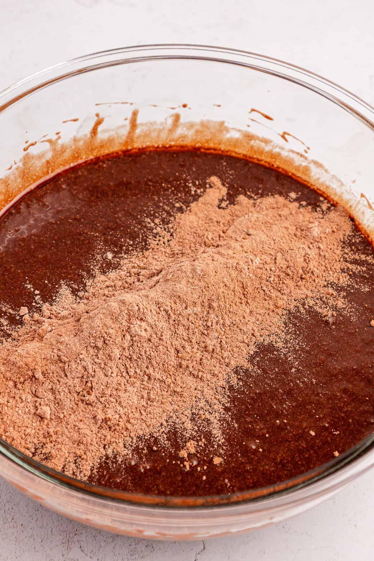 Una ciotola di vetro piena di impasto per brownie al cioccolato, con un mucchio di cacao in polvere secco e farina sopra, parzialmente mischiati.