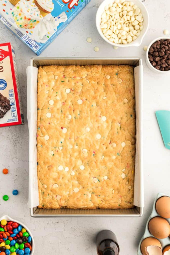Barrette di biscotti da preparato per torte 12 a rectangular pan of funfetti cake bars that haven't been cut yet, surrounded by two boxes of cake mix, eggs, a bottle of vanilla, a bowl of M&Ms, a small bowl of chocolate chips and a larger bowl of white chocolate chips