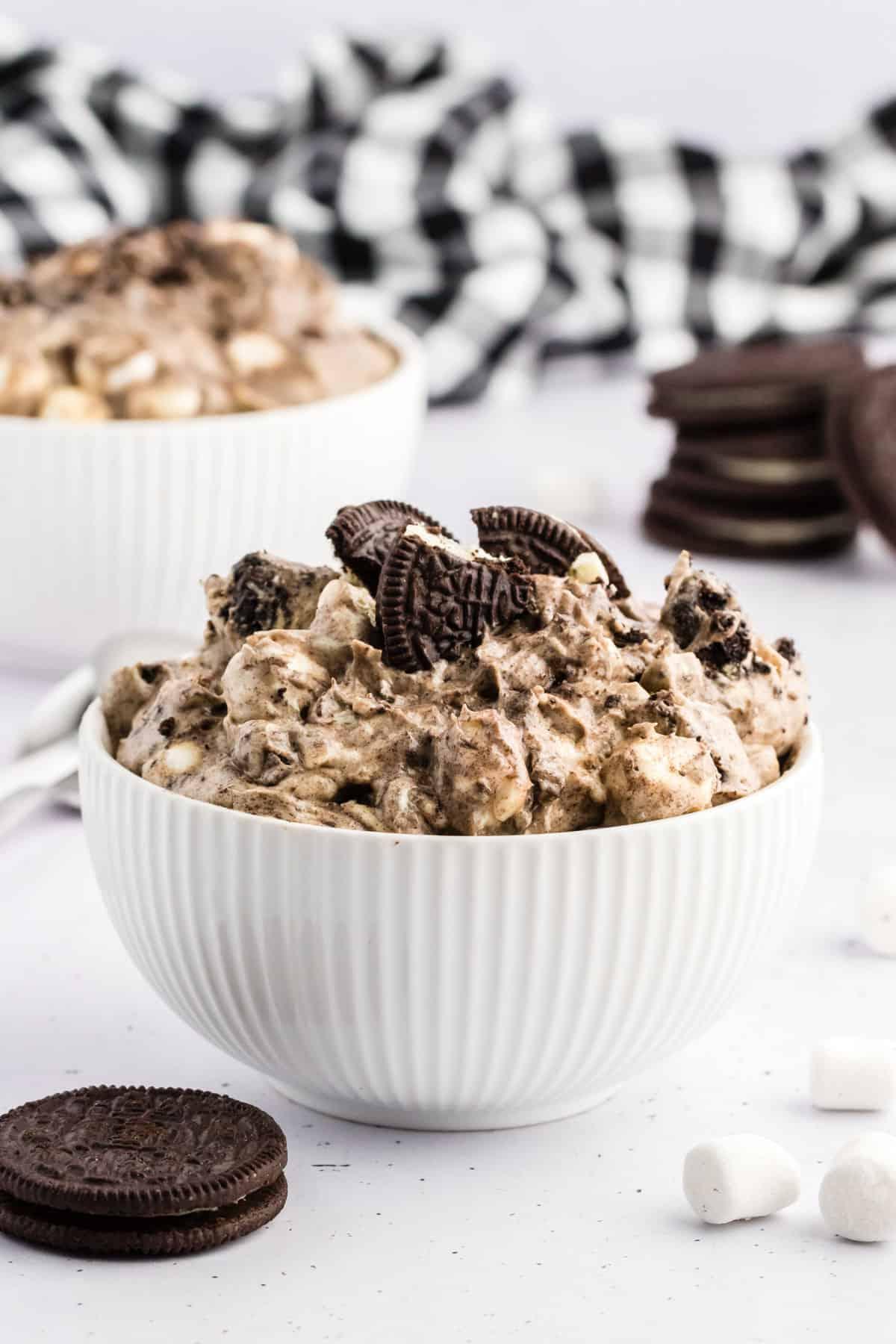 Ricetta Crema Soffice Oreo (Senza Formaggio Cremoso) 12 Fluff di Oreo in una ciotola bianca circondato da biscotti Oreo interi, mini marshmallow e un panno da cucina a scacchi bianchi e neri