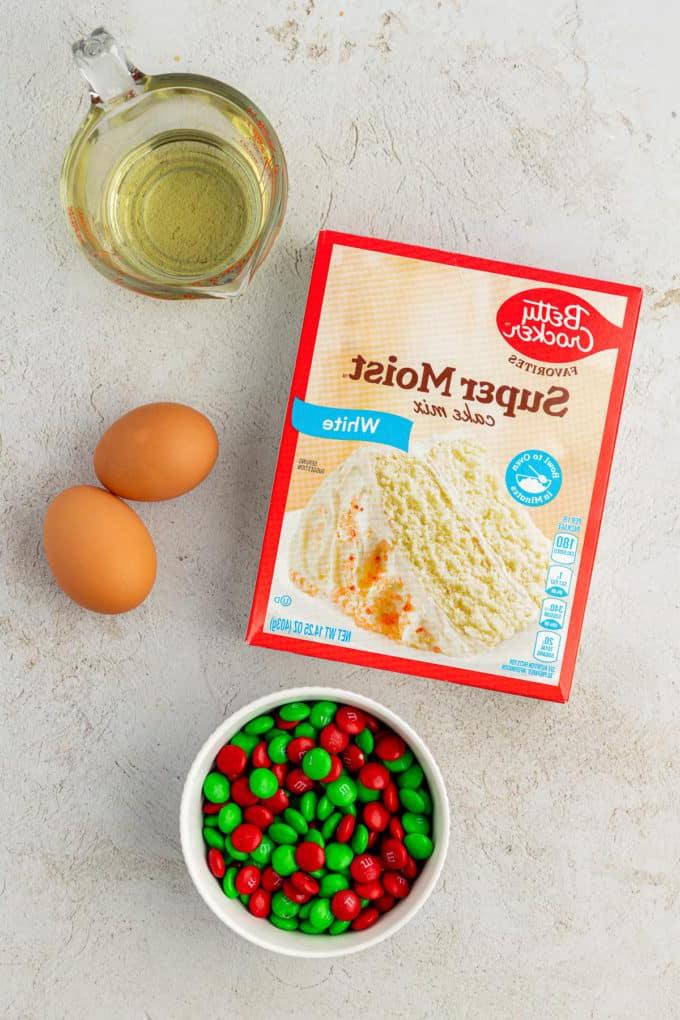 Fare Biscotti dal Mix per Torte 12 Ingredienti per biscotti natalizi con codette e red velvet su un piano di lavoro, inclusi una scatola di preparato per torte bianco, olio vegetale, due uova e una ciotola di M&M's verdi e bianchi