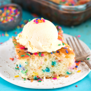 Torta Terremoto Funfetti 2 Una fetta di torta terremoto funfetti guarnita con gelato alla vaniglia e zuccherini colorati, posata su un piatto bianco, con una forchetta e altri zuccherini sullo sfondo.