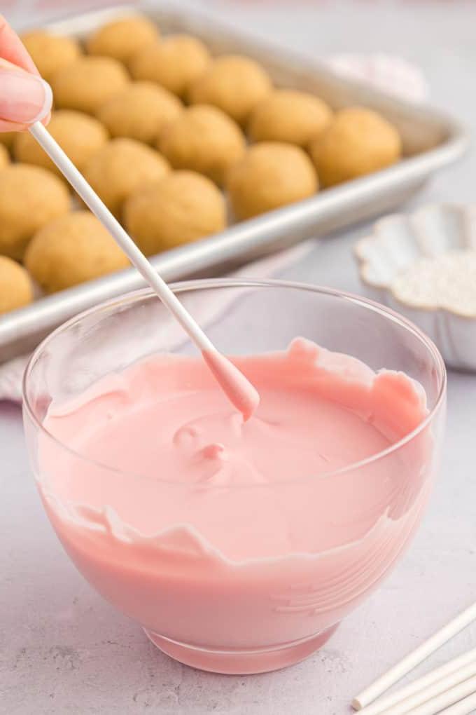 un bastoncino per cake pop appena intinto in cioccolato fuso rosa sopra una ciotola di vetro trasparente di cioccolato fuso, con una teglia di cake pops semplici dietro