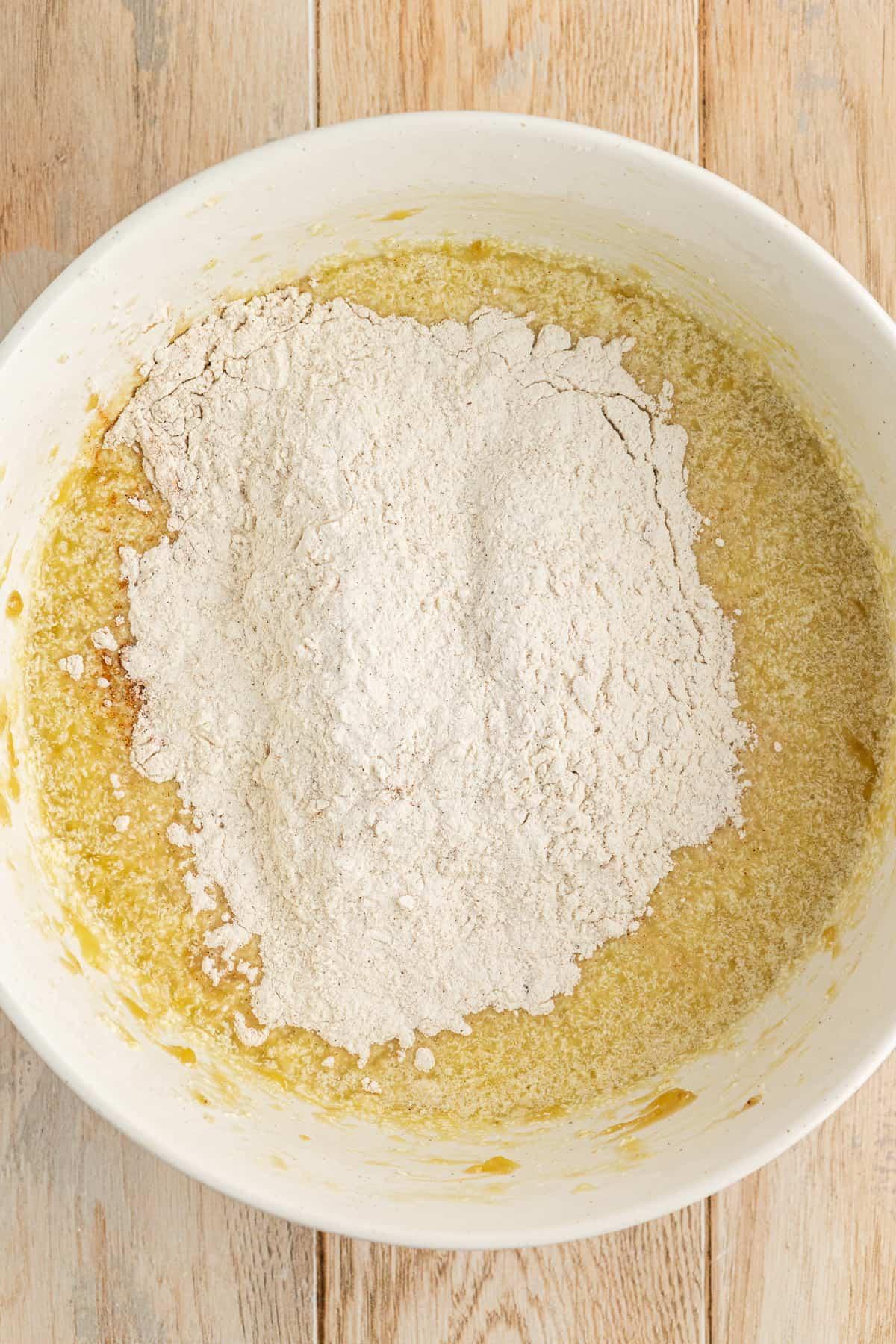 Ricetta Facile Torta alla Banana 12 Una ciotola bianca su una superficie di legno con ingredienti umidi per la torta di banane e una miscela di farina secca sopra non ancora mescolata