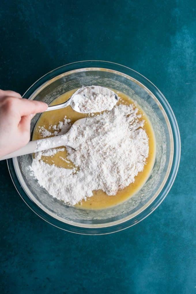 Il Miglior Pan di Banane con Gocce di Cioccolato 12 Ingredienti liquidi per il pane alle banane e gocce di cioccolato in una ciotola di vetro trasparente su una superficie color ottanio scuro con ingredienti secchi che vengono aggiunti sopra.