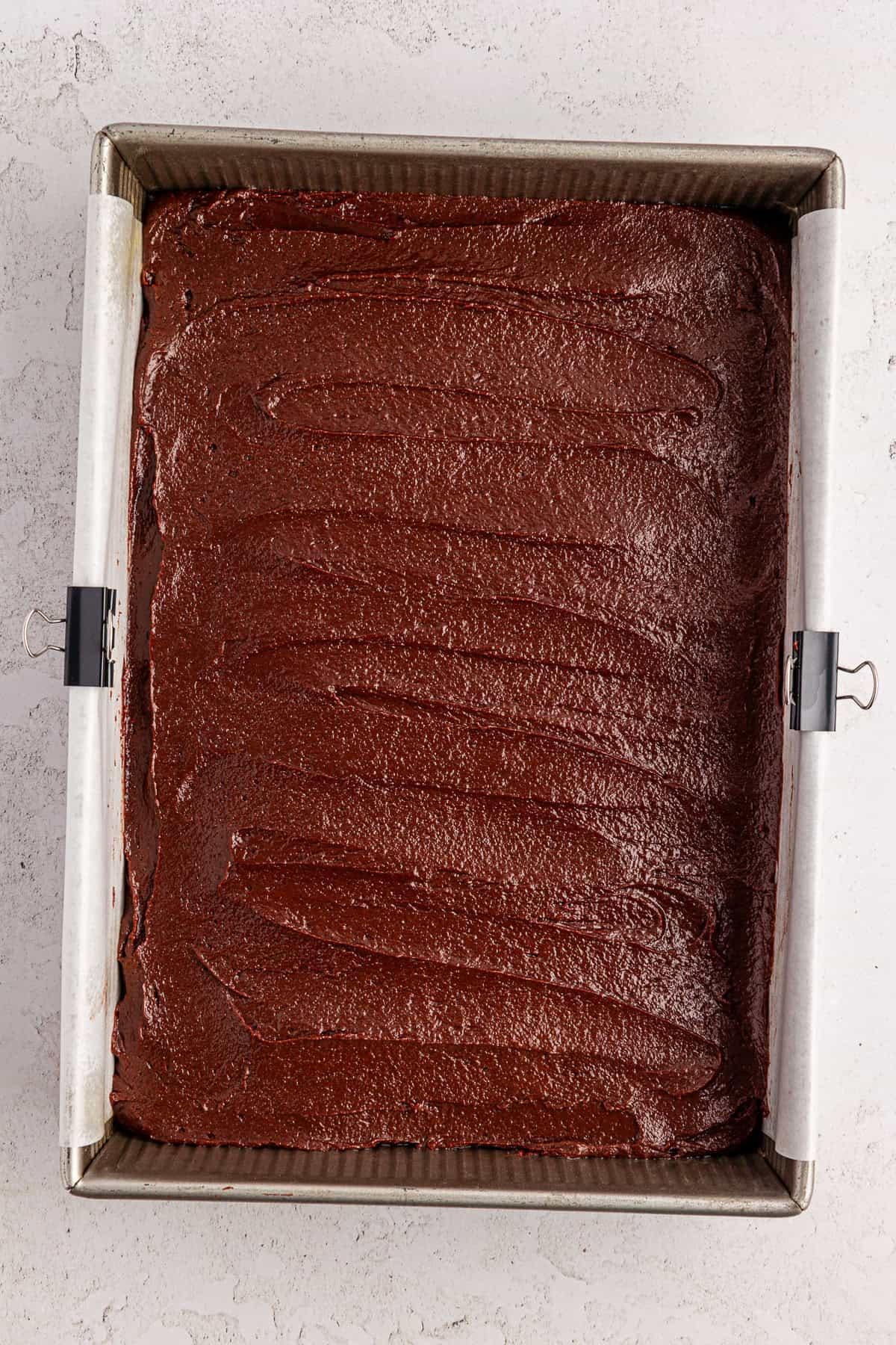 Una teglia da forno quadrata foderata con carta forno contiene uno strato uniforme di impasto per brownie al cioccolato, pronto per essere infornato. L'impasto è lucido e spalmato in modo uniforme con spirali visibili sulla superficie.