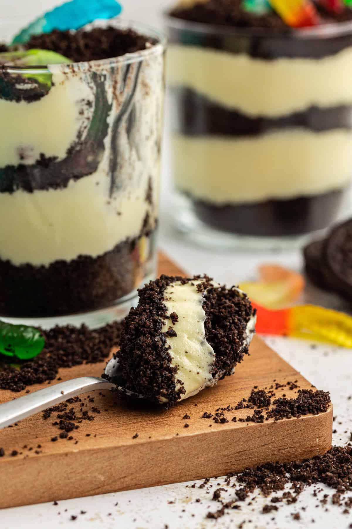 La migliore ricetta per la Torta al Fango 13 Un cucchiaio pieno di torta di terra poggiato su un piatto di legno accanto a piccole coppe da trifle di torta di terra con briciole di Oreo e vermi gommosi sparsi intorno