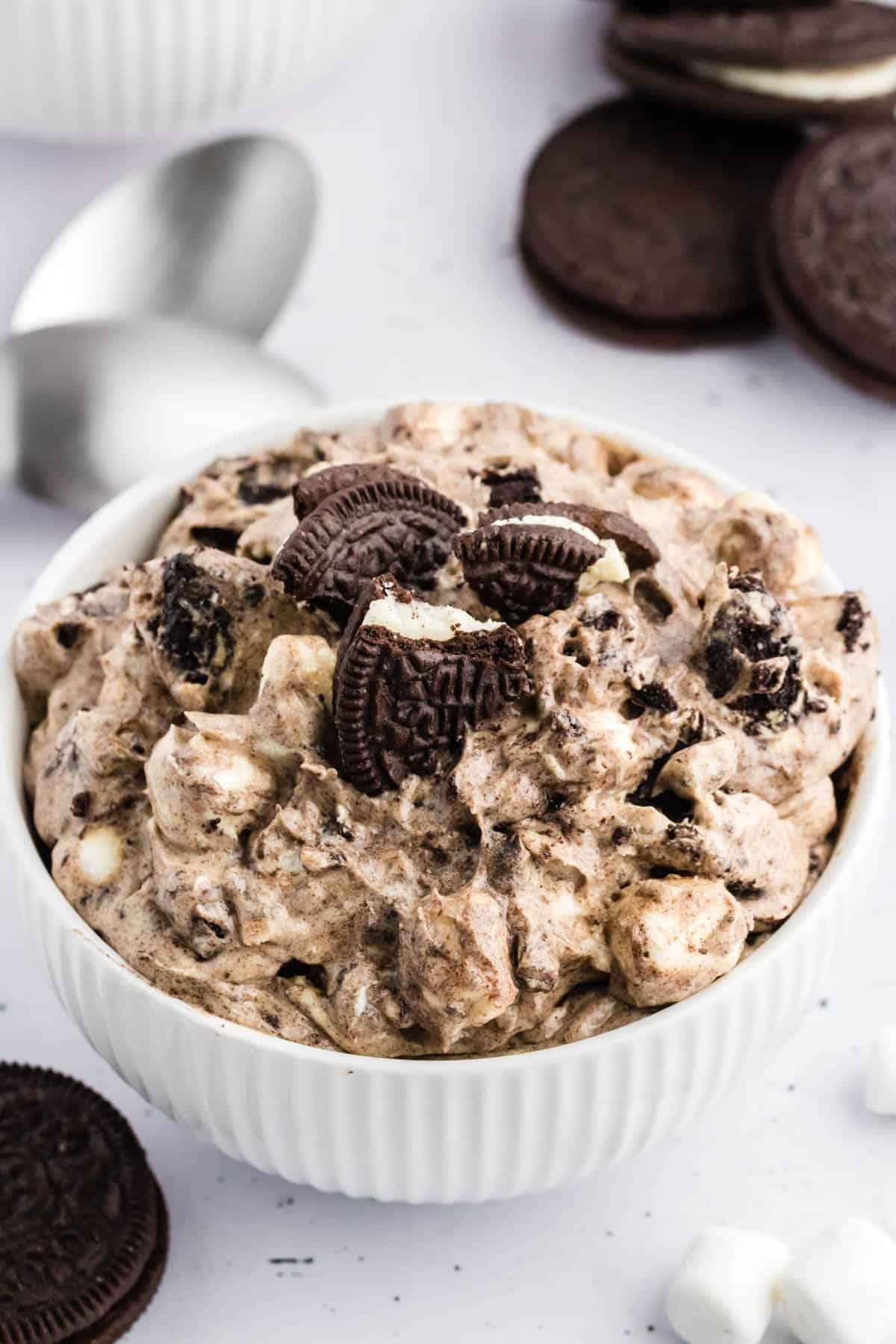 Ricetta Crema Soffice Oreo (Senza Formaggio Cremoso) 13 Fluff di Oreo in una ciotola bianca circondato da biscotti Oreo interi, mini marshmallow e un panno da cucina a scacchi bianchi e neri