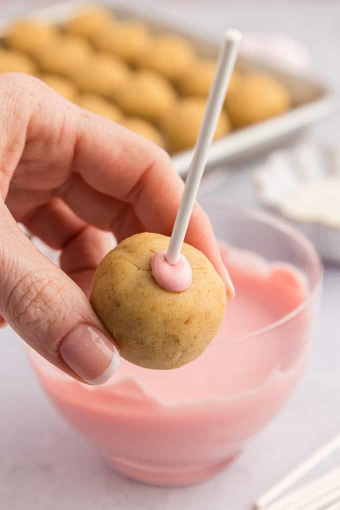 una mano che tiene un cake pop su un bastoncino con un po' di cioccolato fuso rosa intorno al punto in cui il bastoncino è entrato nel cake pop, con una ciotola di cioccolato fuso rosa e una teglia di cake pops semplici sullo sfondo