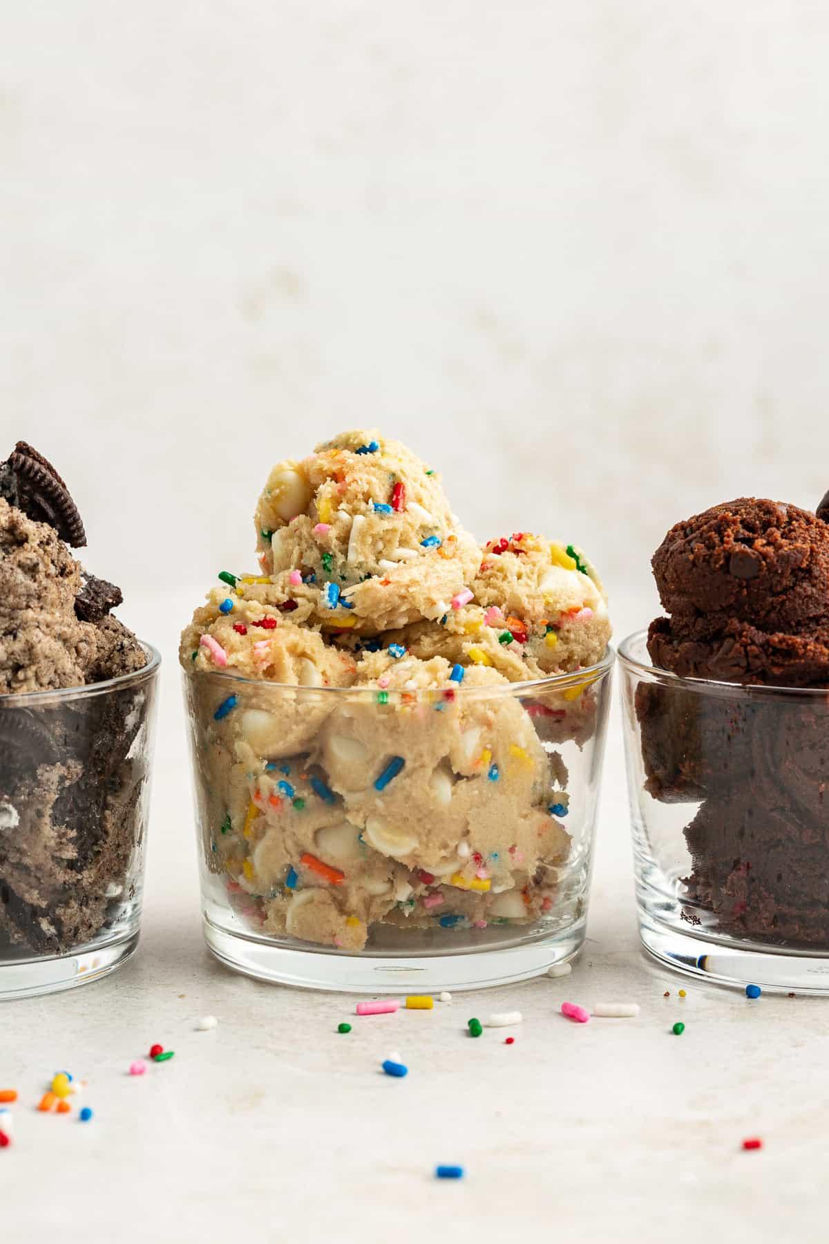 **Semplice Impasto per Biscotti Commestibile** 13 three round glass containers full of edible cookie dough with chocolate on the left, funfetti in the middle and oreo flavor on the right, with rainbow sprinkles scattered around