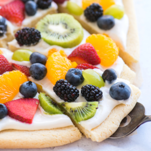 Primo piano di una deliziosa fetta di pizza di frutta guarnita con glassa al formaggio cremoso, mirtilli, more, kiwi, fragole, uva verde e spicchi d'arancia su una morbida base di biscotto zuccherato.