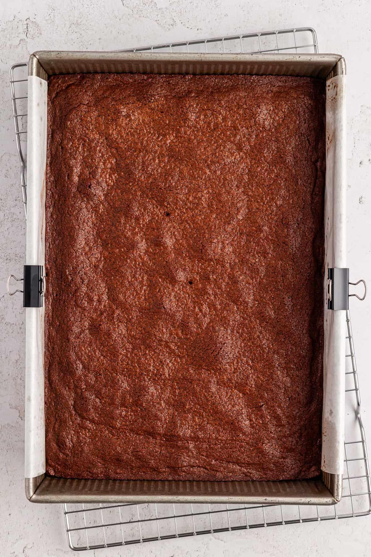 Una teglia quadrata di brownies appena sfornati, foderata con carta forno e fissata ai lati, è posta su una griglia di raffreddamento metallica sopra una superficie bianca.