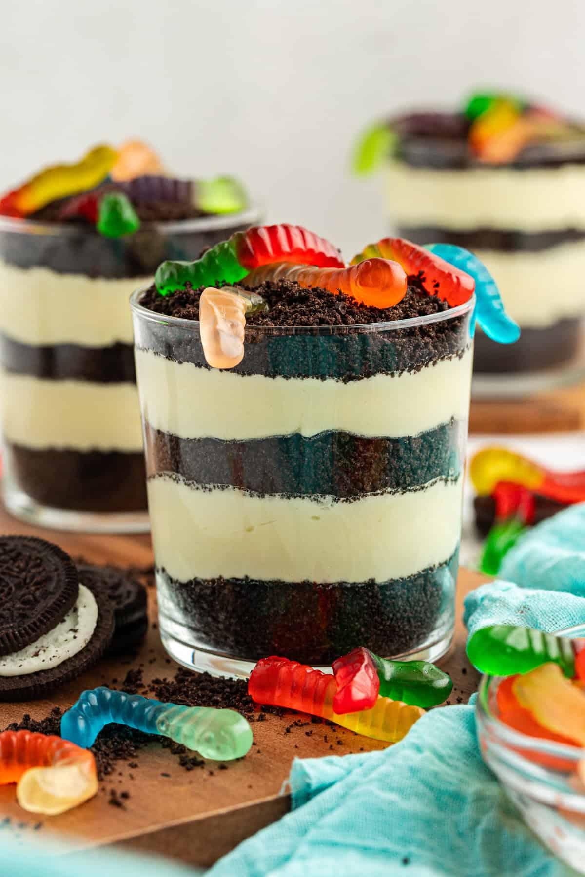 La migliore ricetta per la Torta al Fango 14 Tre piccole coppe da trifle di torta di terra guarnite con vermi gommosi su un piatto di legno circondate da Oreo, Oreo sbriciolati, vermi gommosi e un asciugamano color ottanio chiaro