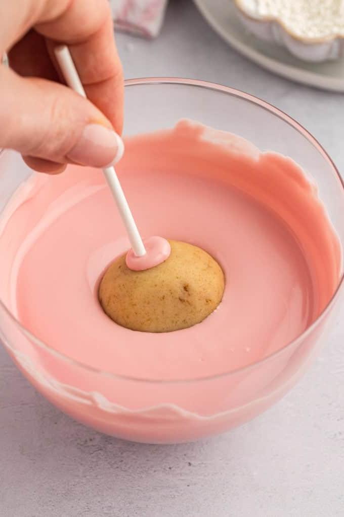un cake pop su un bastoncino bianco che viene intinto in una ciotola di vetro trasparente piena di cioccolato fuso rosa