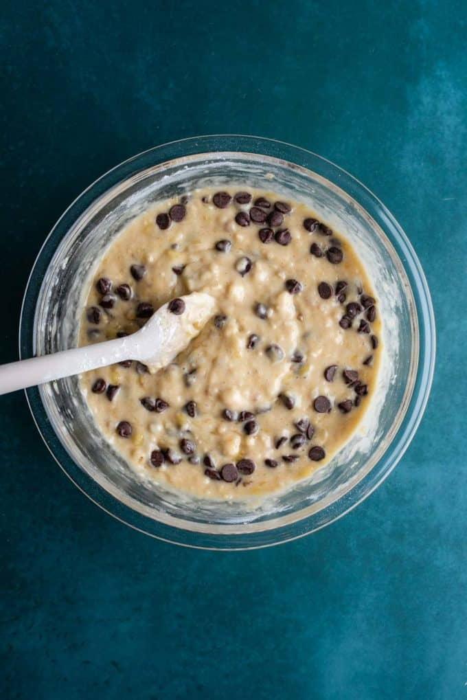 Il Miglior Pan di Banane con Gocce di Cioccolato 14 Impasto del pane alle banane e gocce di cioccolato mescolato in una ciotola di vetro trasparente con una spatola bianca su una superficie color ottanio scuro.