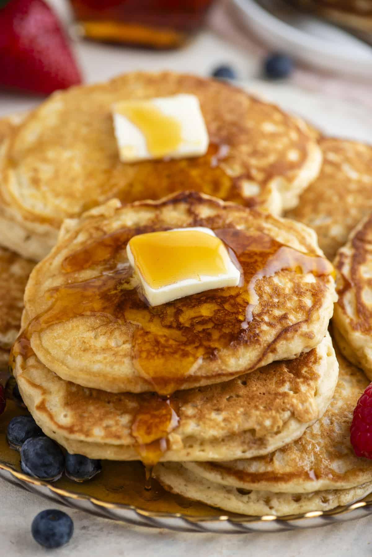 Pancakes Soffici Fatti in Casa 14 Pancake impilati su un piatto, sormontati da burro e conditi con sciroppo, circondati da frutti di bosco freschi, con altri frutti di bosco e pancake sullo sfondo.